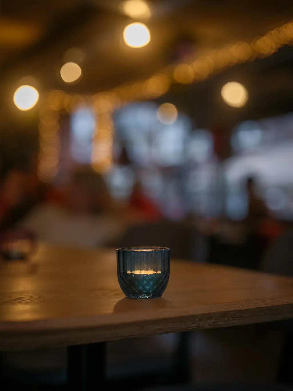 A small glass candle holder glowing on a wooden table, with warm lights blurred across the room behind.