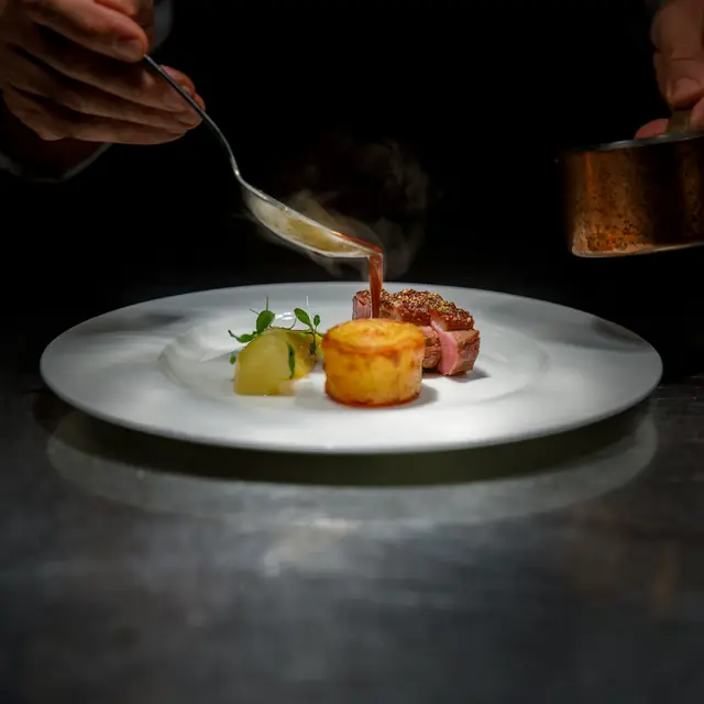 A cook spooning sauce over a plated dish of meat and potato at the pass.