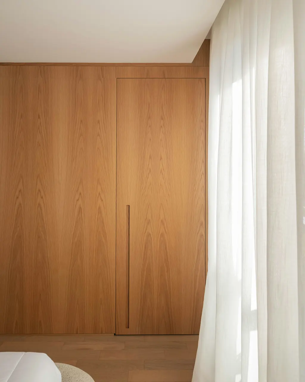 A fitted oak wardrobe beside a window with a sheer curtain.