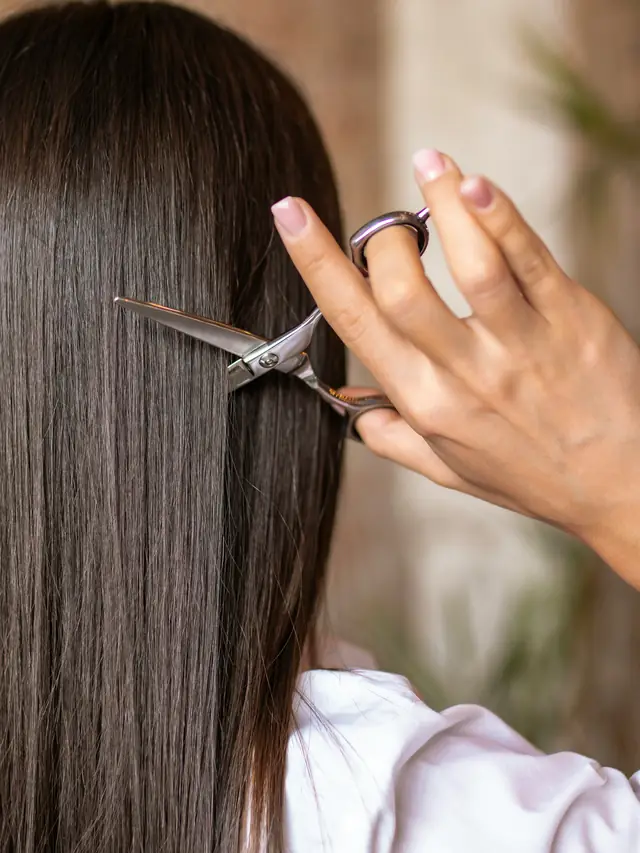 A stylist’s hands trimming the ends of long, straight dark hair with scissors.