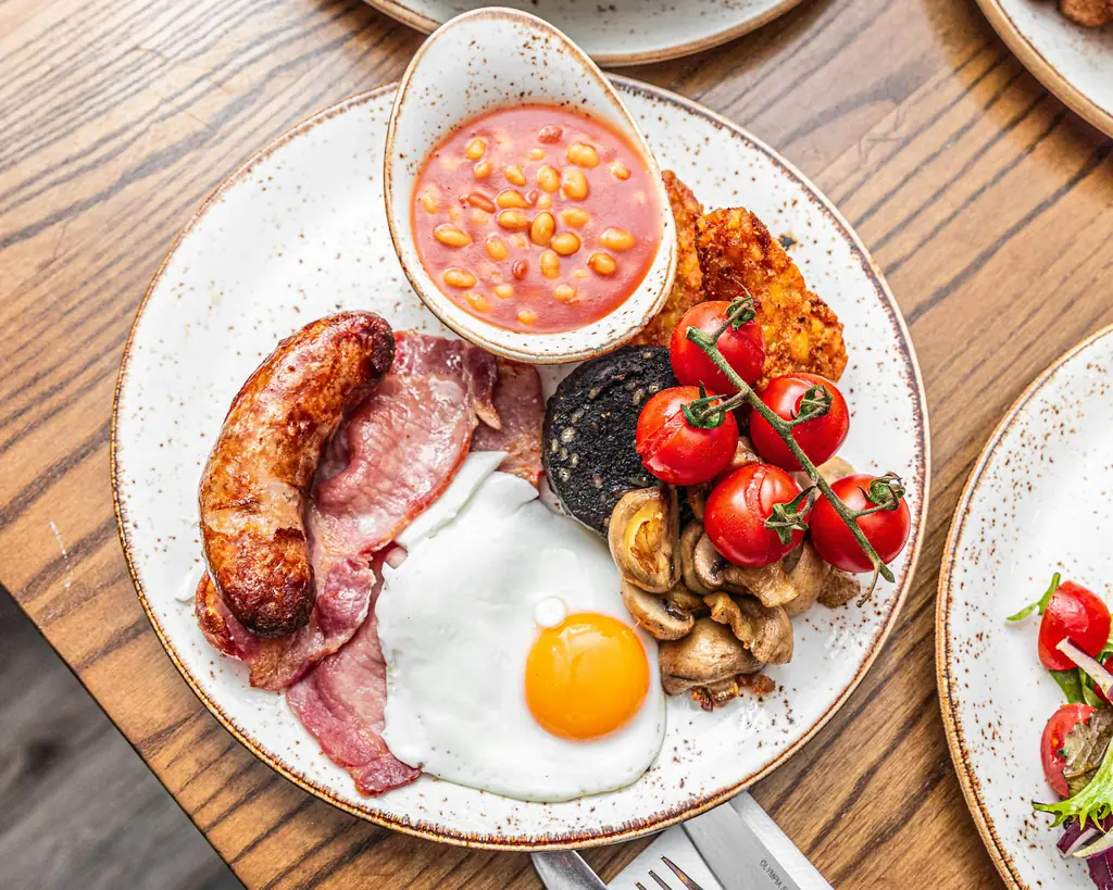 A cooked breakfast on a plate: sausage, bacon, a fried egg, mushrooms, tomatoes on the vine and a pot of beans, on a wooden table.