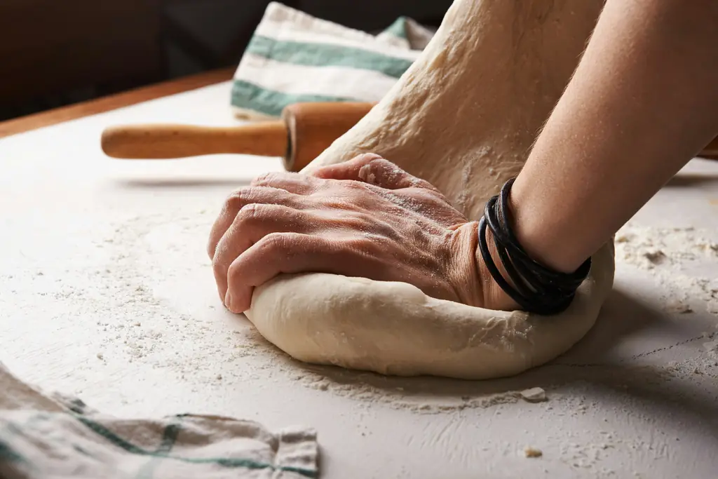 A baker’s hands shaping a round of dough on a floured wooden bench, a rolling pin beside them.