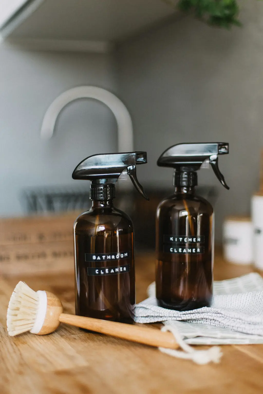 Two amber glass spray bottles labelled bathroom cleaner and kitchen cleaner, with a wooden scrubbing brush, on a kitchen worktop.
