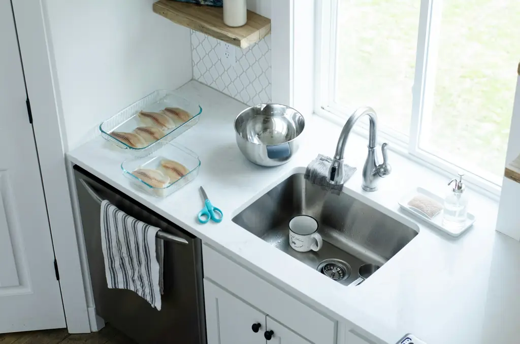 A bright kitchen corner: a steel sink under a window, a clean white worktop and a tea towel on the dishwasher door.