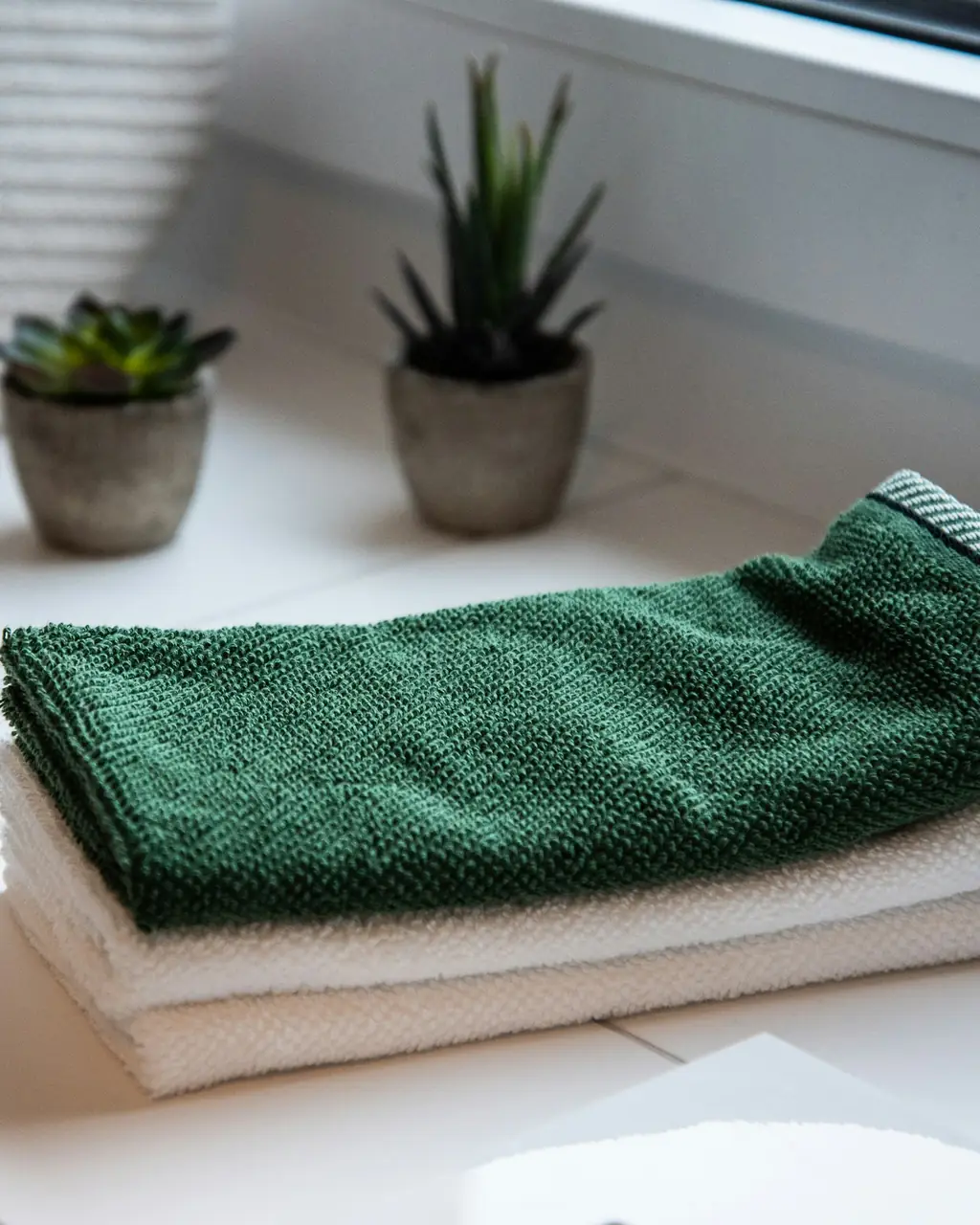 Folded green and white cloths on a clean white windowsill, beside two small potted plants.