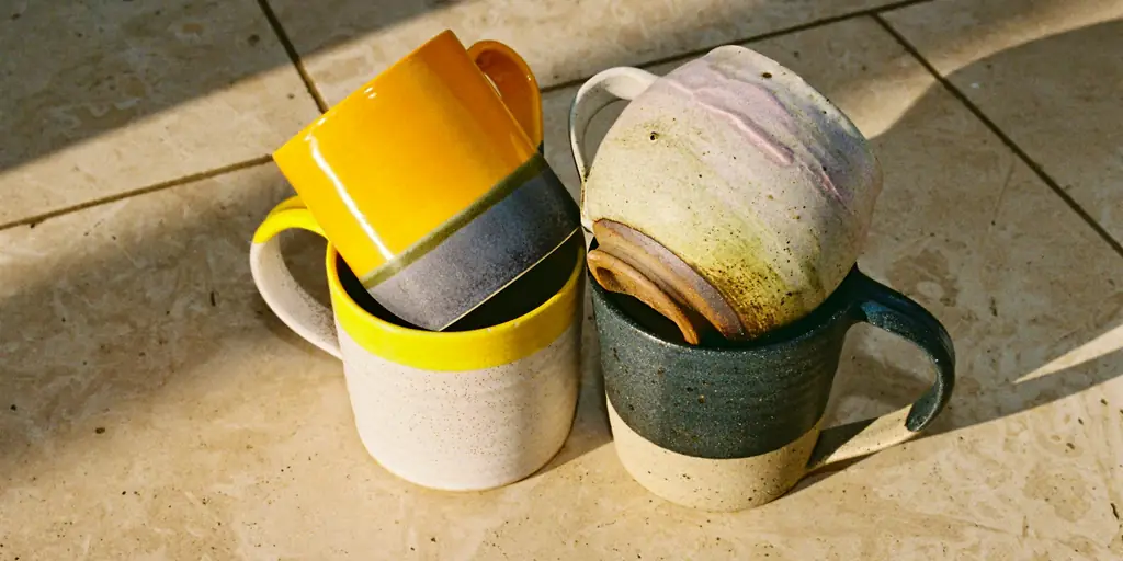 Four stoneware mugs stacked in pairs on a sunny stone floor: one in a bright marigold glaze, one rimmed in yellow, one speckled lilac and one dark blue.