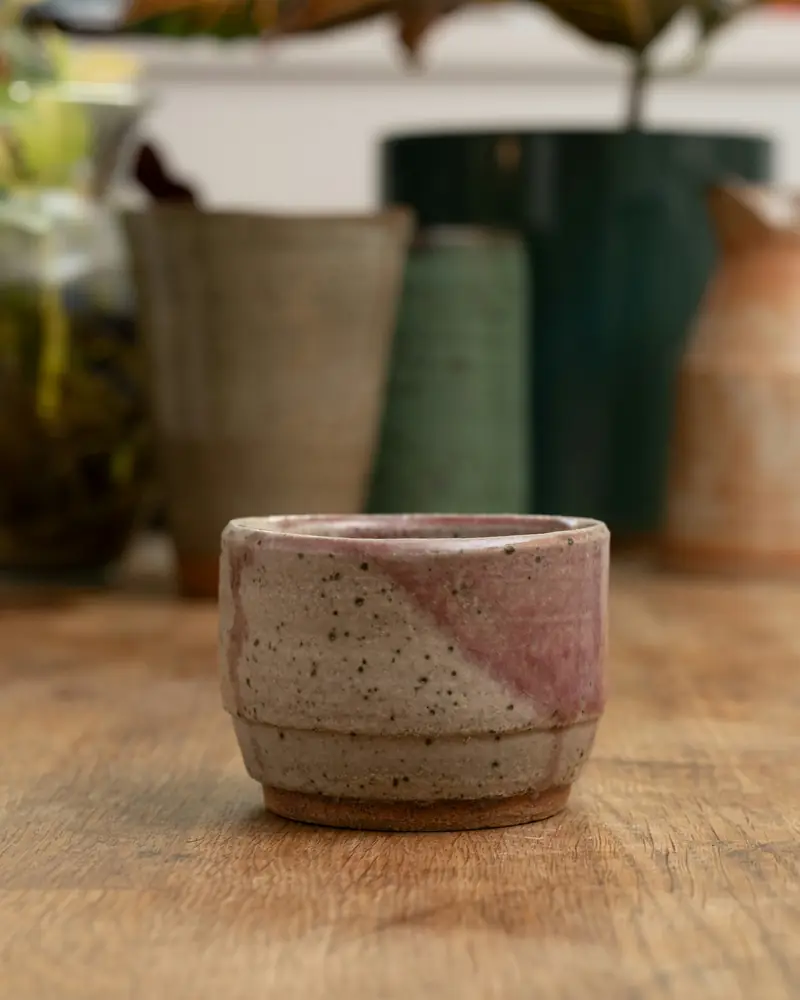 A small speckled cup dipped at an angle in a soft pink glaze, on a wooden table.