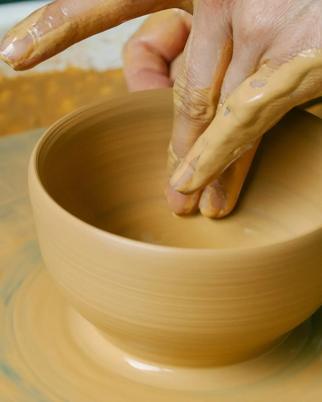 Two hands shaping a bowl of pale yellow clay on a spinning potter’s wheel.