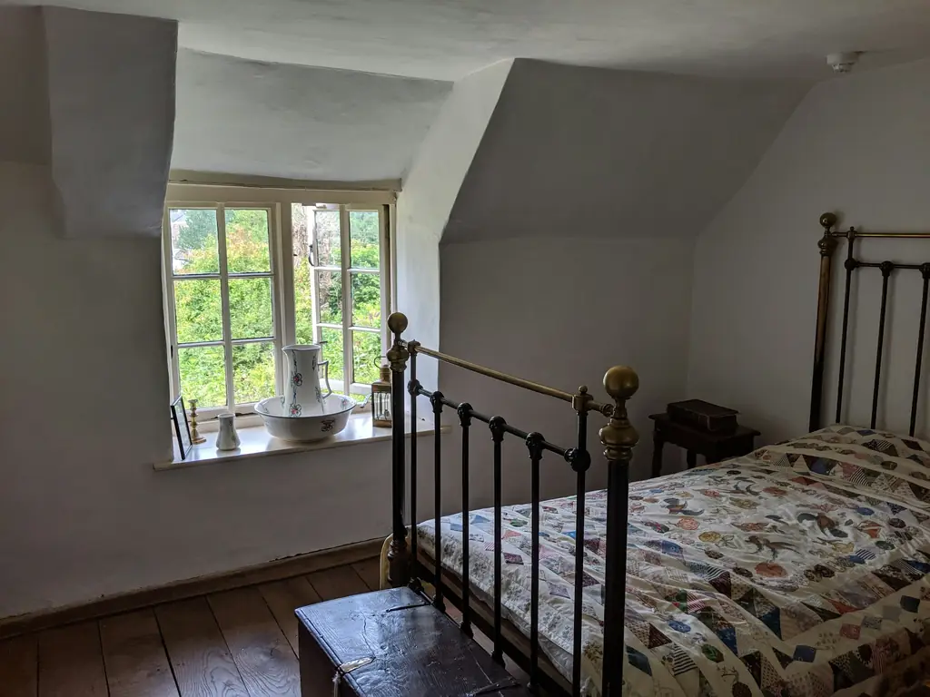A whitewashed bedroom under sloping eaves: a brass and iron bed with a patchwork quilt, and a jug and basin on the window sill.