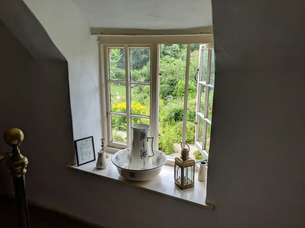 A deep window sill in a thick whitewashed wall, with a jug, a basin and a lantern, and a green garden through the open window.