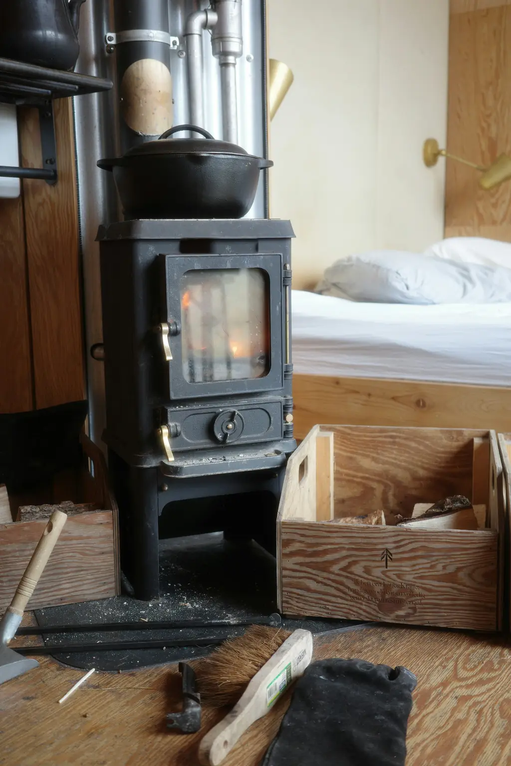 A small cast-iron wood-burner with a pot on top, beside a bed made up in white linen and a box of kindling.