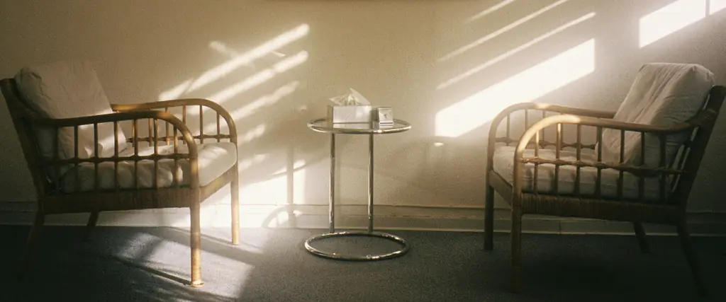 Two wooden armchairs either side of a small glass table, with stripes of morning sun across the wall and floor.