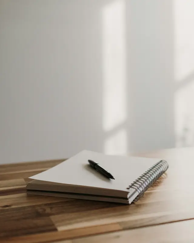 A closed notebook with a pen on a wooden table, lit by a tall window.
